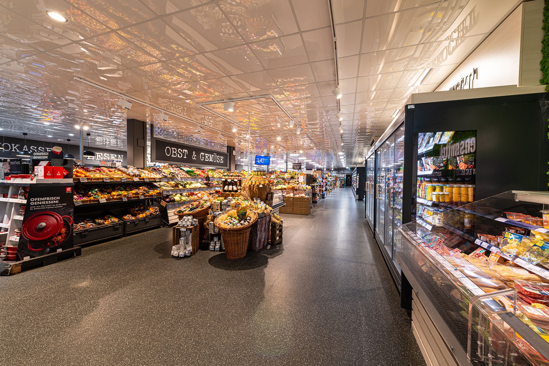 Ein gut beleuchteter Supermarktgang mit Regalen voller Obst, Gemüse und gekühlter Lebensmittel; die Beschilderung „Obst & Gemüse“ ist sichtbar.