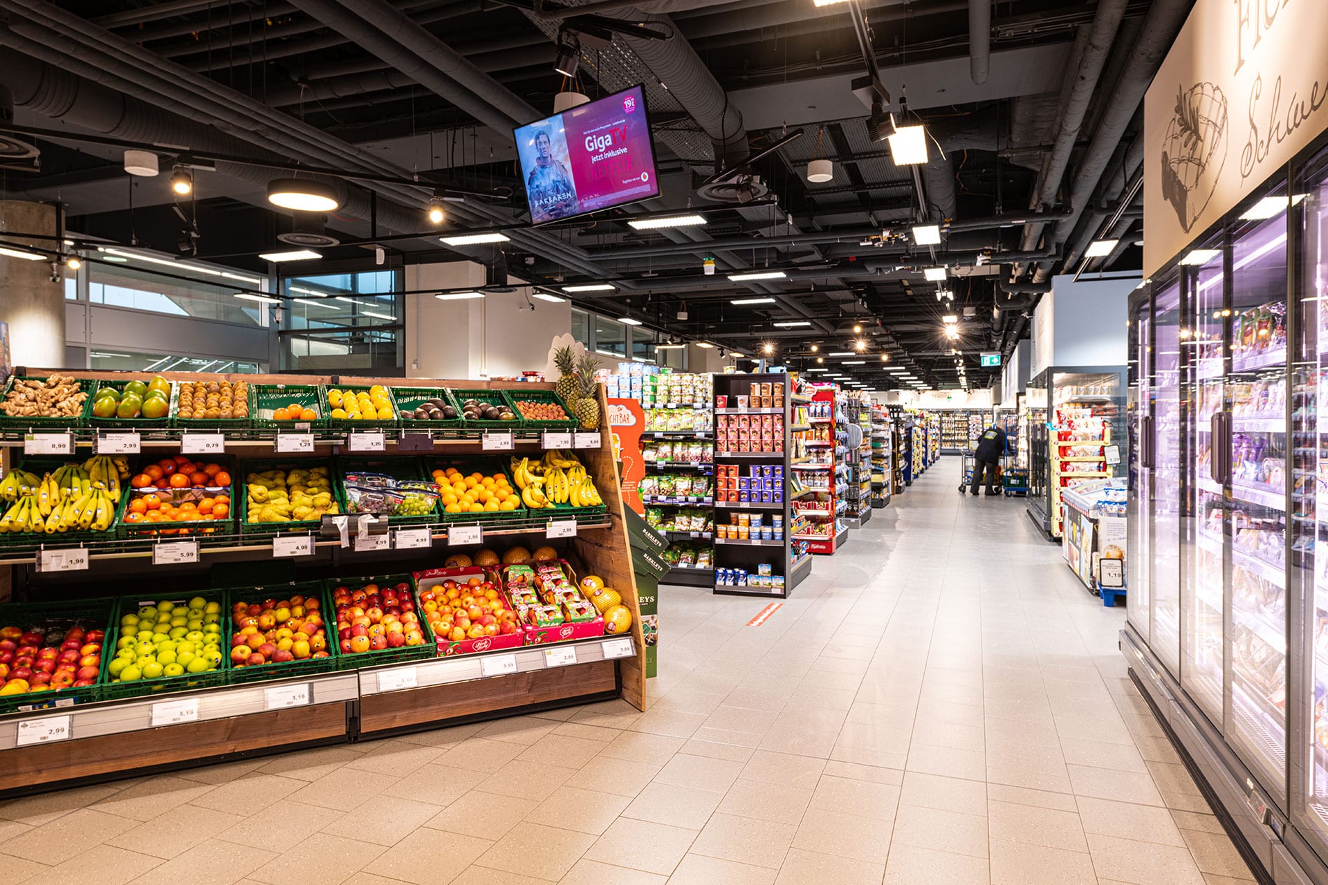 Ein hell erleuchteter Supermarktinnenraum mit frischen Produkten auf der linken Seite, gekühlten Waren auf der rechten Seite und Lebensmittelgängen, die sich nach hinten erstrecken.