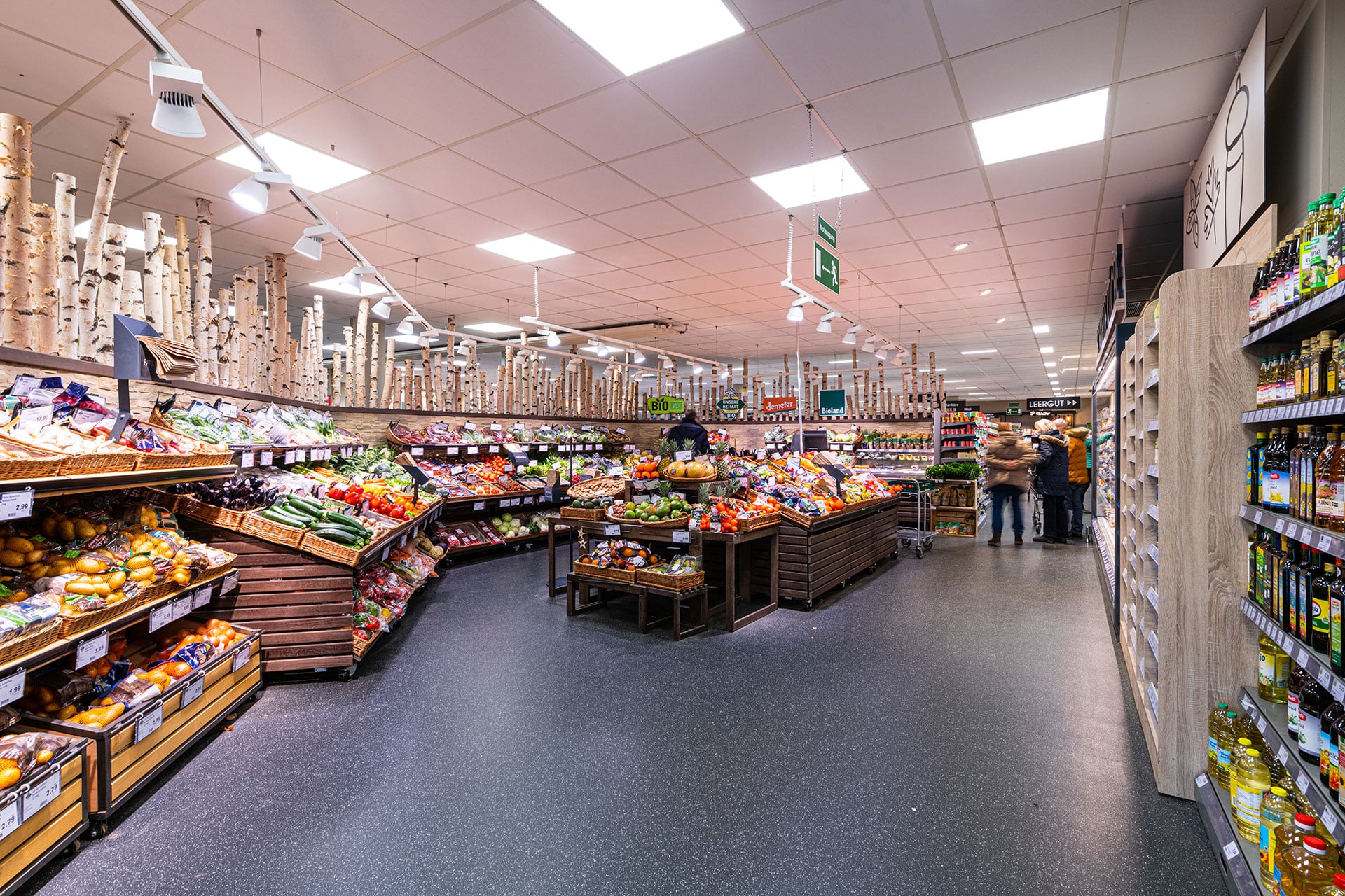 Die Obst- und Gemüseabteilung eines Lebensmittelladens mit verschiedenen Obst- und Gemüsesorten auf Holzständern, Regalen mit verpackten Waren auf der rechten Seite und Käufern im Hintergrund.