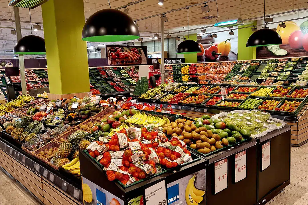 In der Obst- und Gemüseabteilung eines Supermarkts wird eine Vielfalt an frischem Obst und Gemüse präsentiert, die auf schrägen Regalen unter schwarzen Hängelampen angeordnet sind.